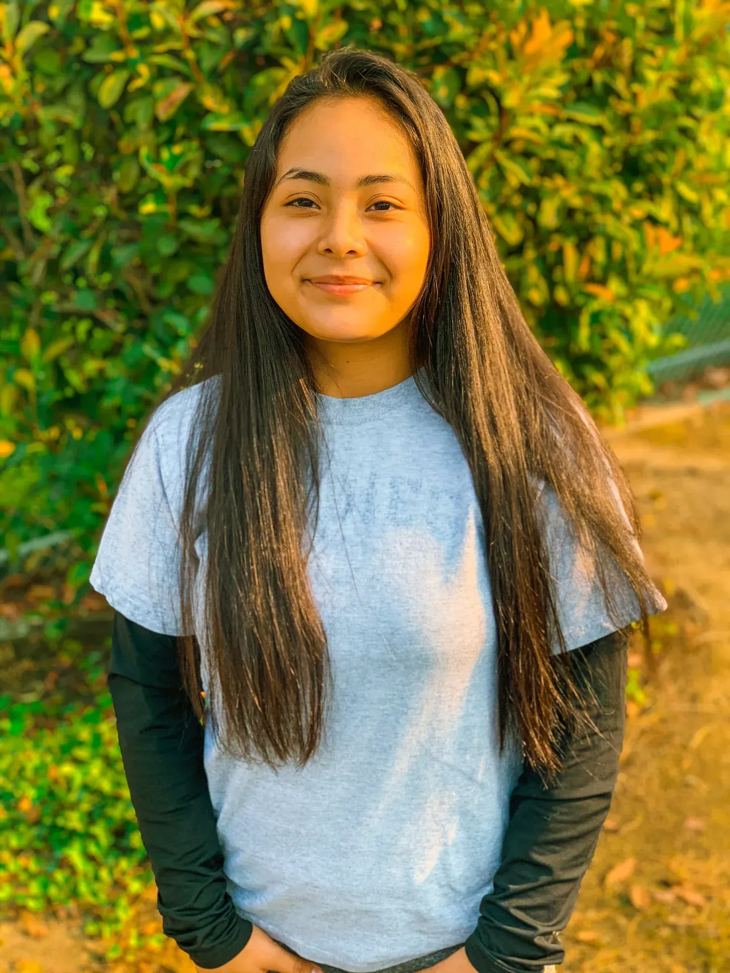 Woman with long dark hair, wearing a gray t-shirt over a black long-sleeve shirt, smiles outdoors.