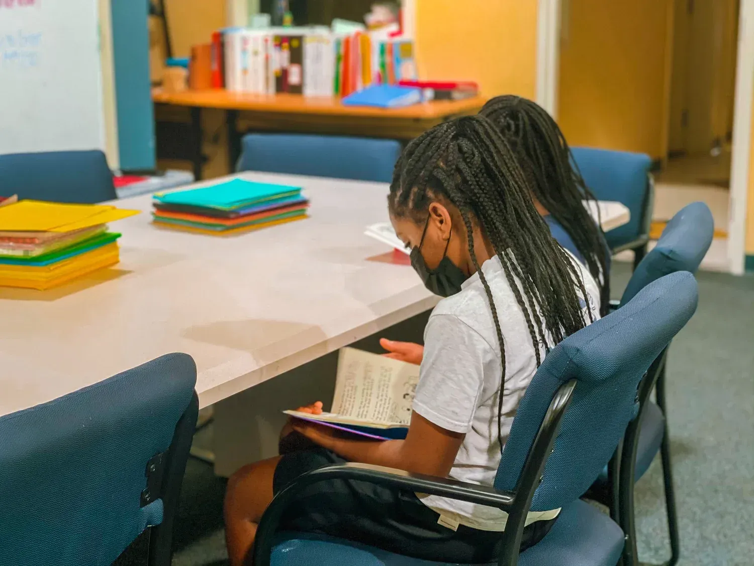 Two people read at a table, books and papers nearby. Blue chairs, white table.