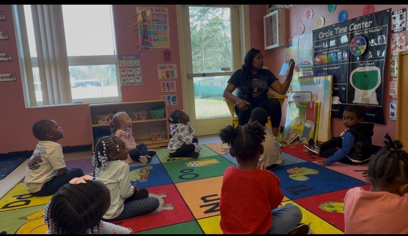 A teacher sits in a chair reading a book to a group of children sitting on a colorful carpet in a classroom.