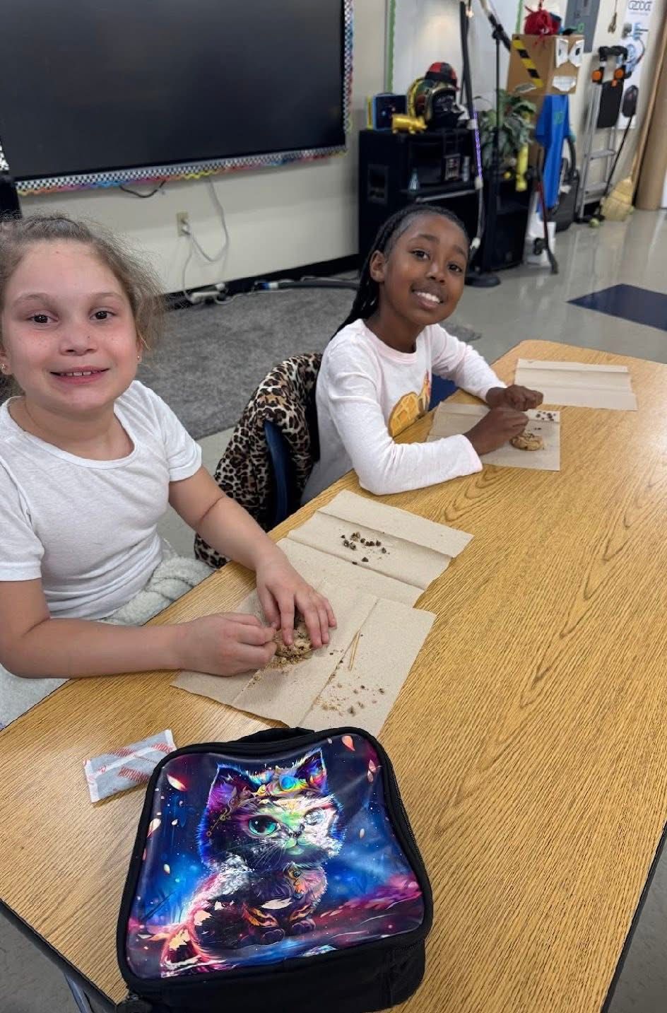 Two students smiling while working with small beads on paper mats at a classroom desk, with a cat-print lunchbox in front.