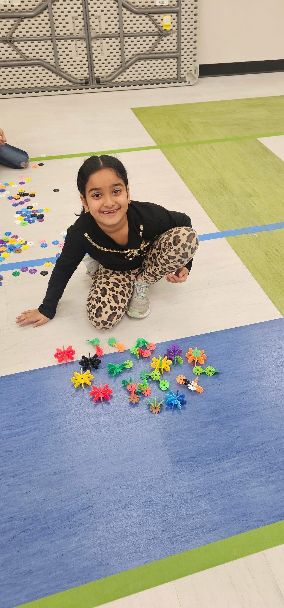 A smiling child kneels on a colorful play mat, surrounded by small, scattered plastic toys.