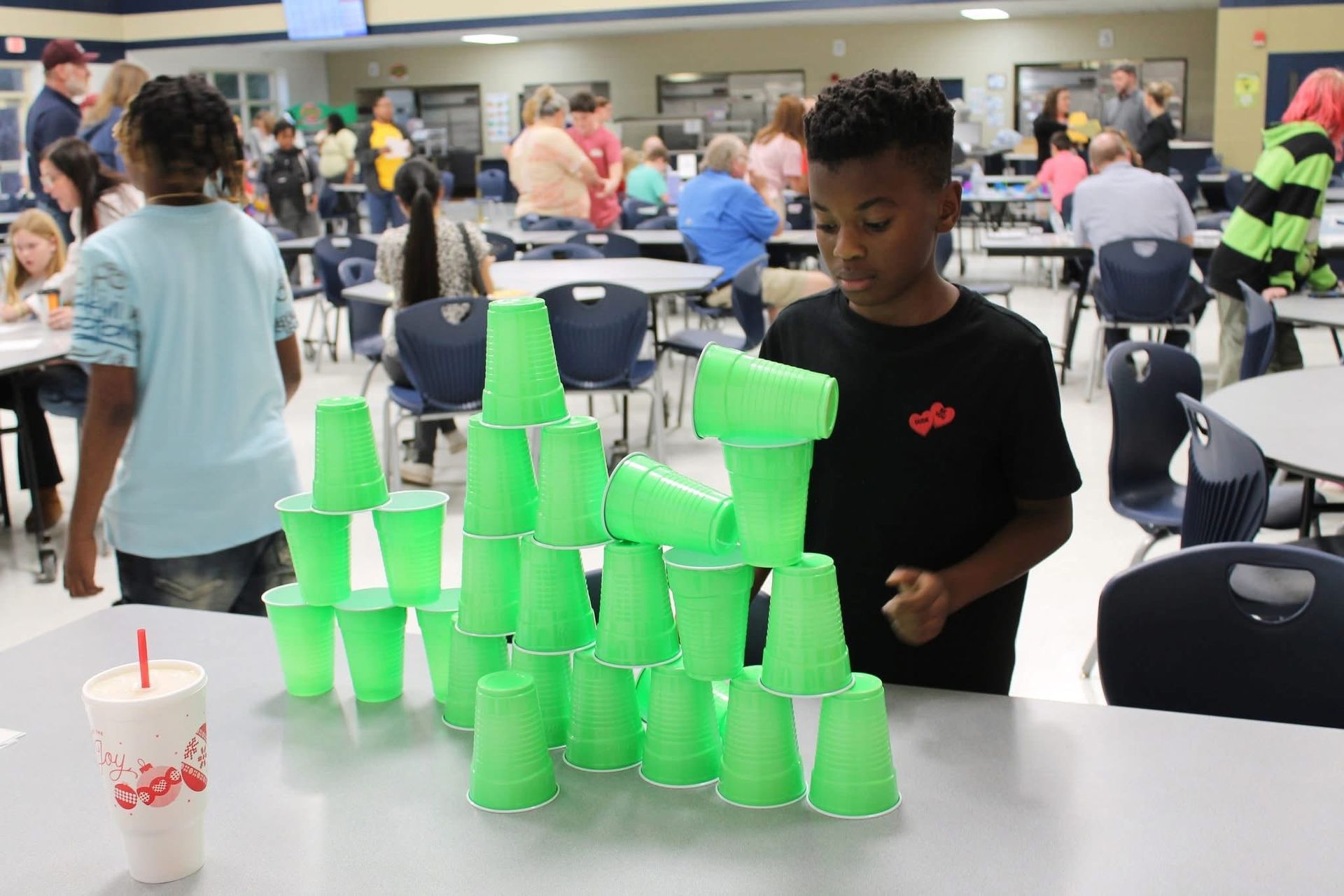 A child builds a tower with green plastic cups on a table in a school cafeteria, with other people in the background.