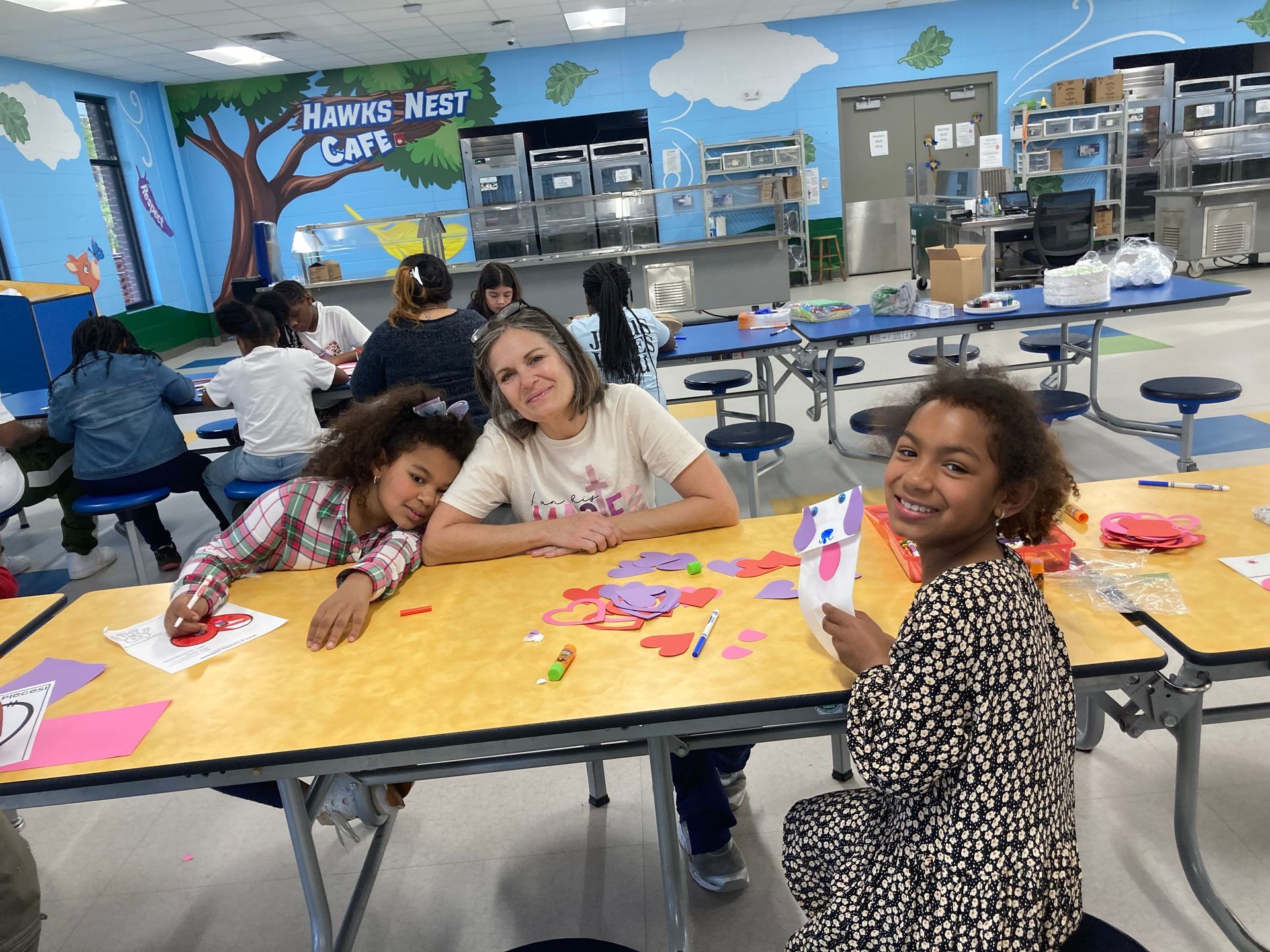 Three children at a yellow cafeteria table creating colorful paper crafts in a room with a mural labeled