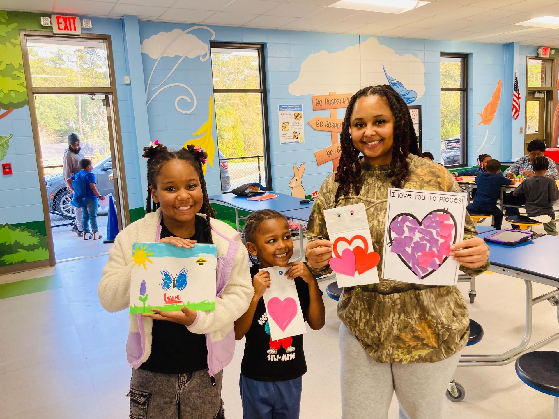 Three people stand in a room, smiling and holding up handmade drawings with heart designs.