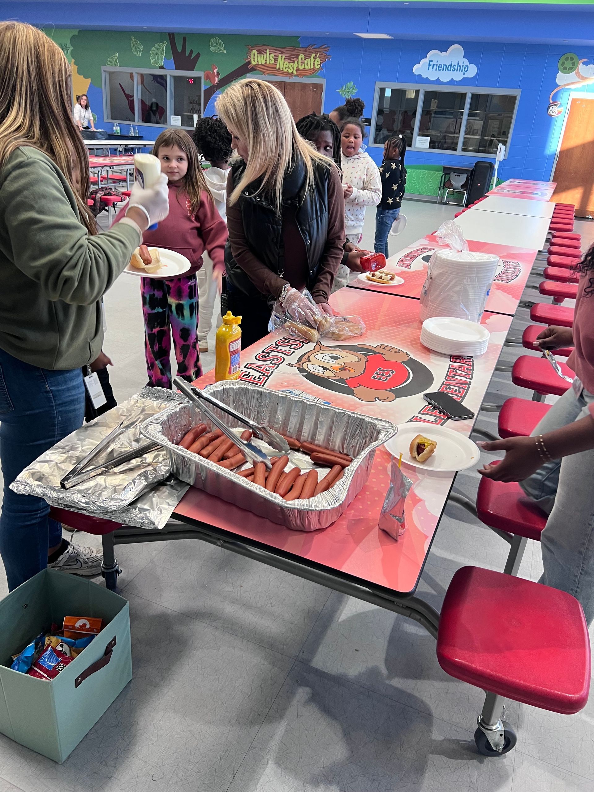 People serve hot dogs from a large aluminum tray on a long table in a school cafeteria decorated with colorful murals.