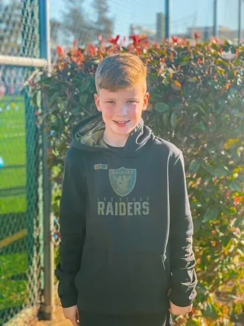Boy in black Raiders hoodie, standing outdoors, smiling, with a soccer field in the background.