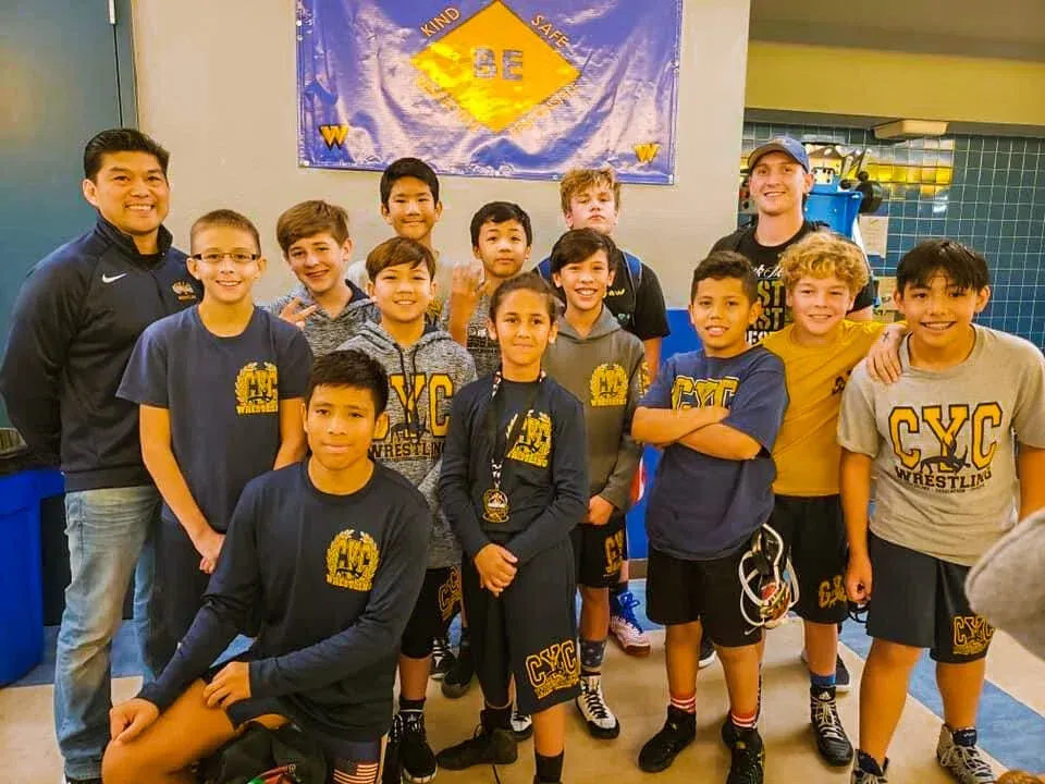 Youth wrestling team posing with coaches in front of a banner. They wear team shirts and look at the camera.