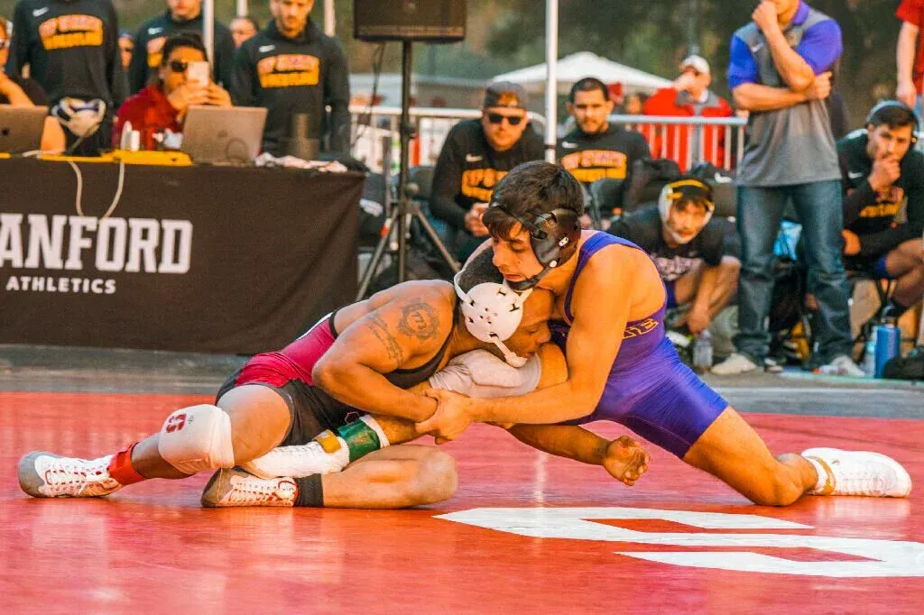 Two wrestlers grappling on a red mat during a Stanford Athletics event. One in purple, one in red.