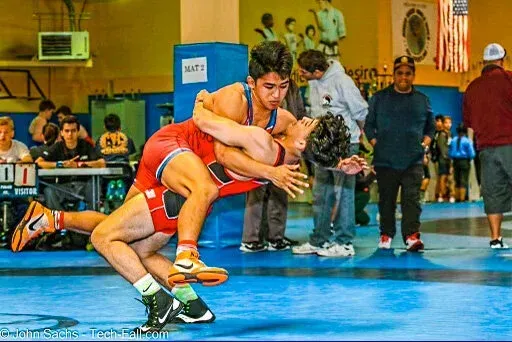Wrestlers grappling on a blue mat. One in red gear has the other in a hold. Spectators watch.