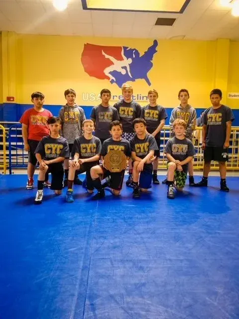Wrestlers in gray and black team shirts pose on a blue mat, holding a championship belt. USA wrestling logo on wall.
