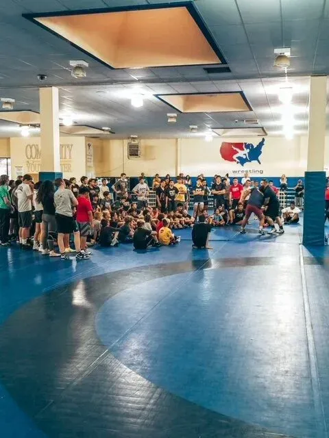 Wrestling practice in a gym: Wrestlers on blue mat, crowd watching. USA wrestling logo on wall.