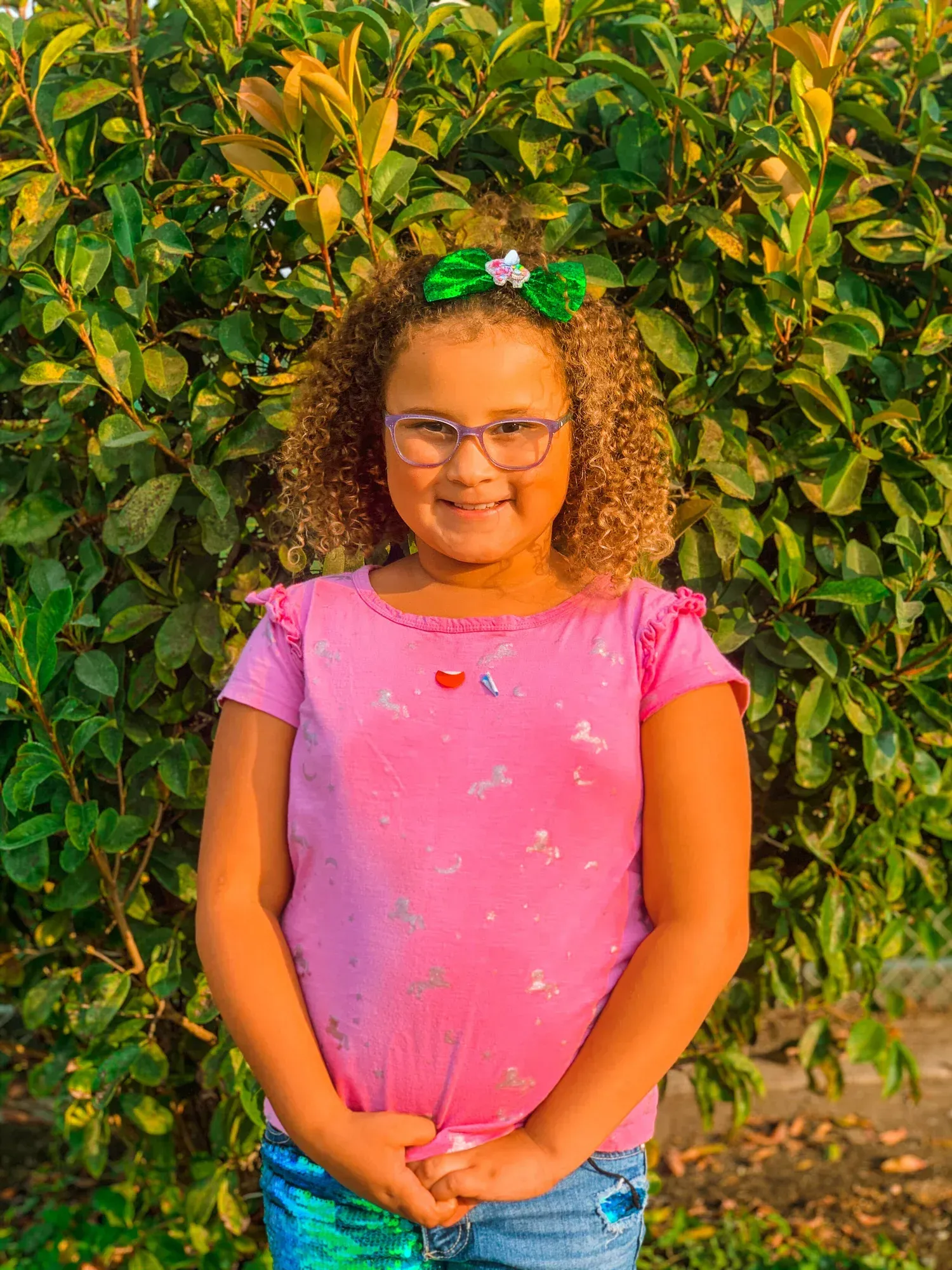 Girl with curly hair wearing glasses, pink shirt, bow, and blue jeans, smiling in front of green bushes.