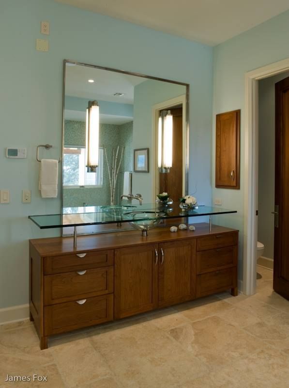 Wooden vanity with glass countertop and mirror in a light blue bathroom, with adjoining doorway visible.