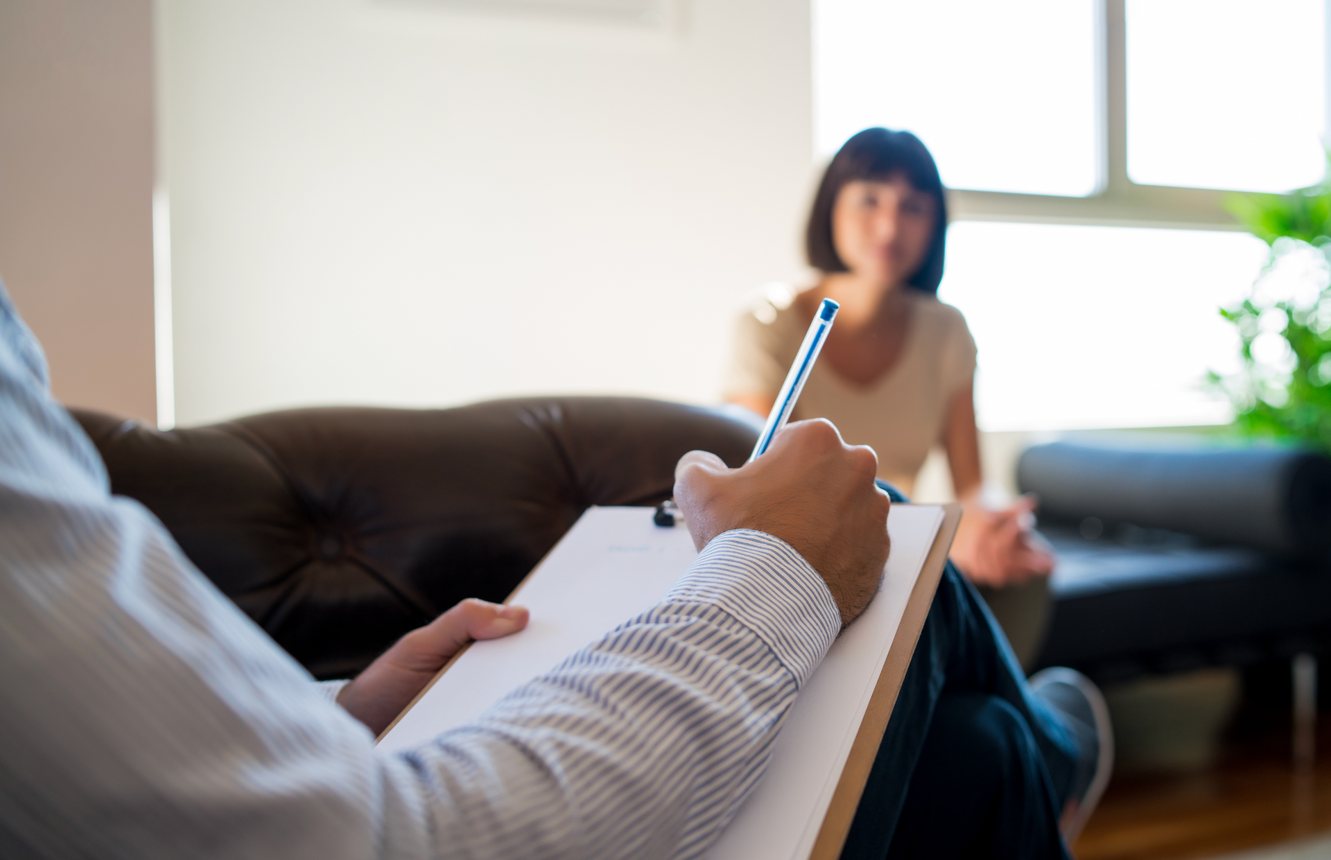 A man is writing on a clipboard while a woman sits on a couch.