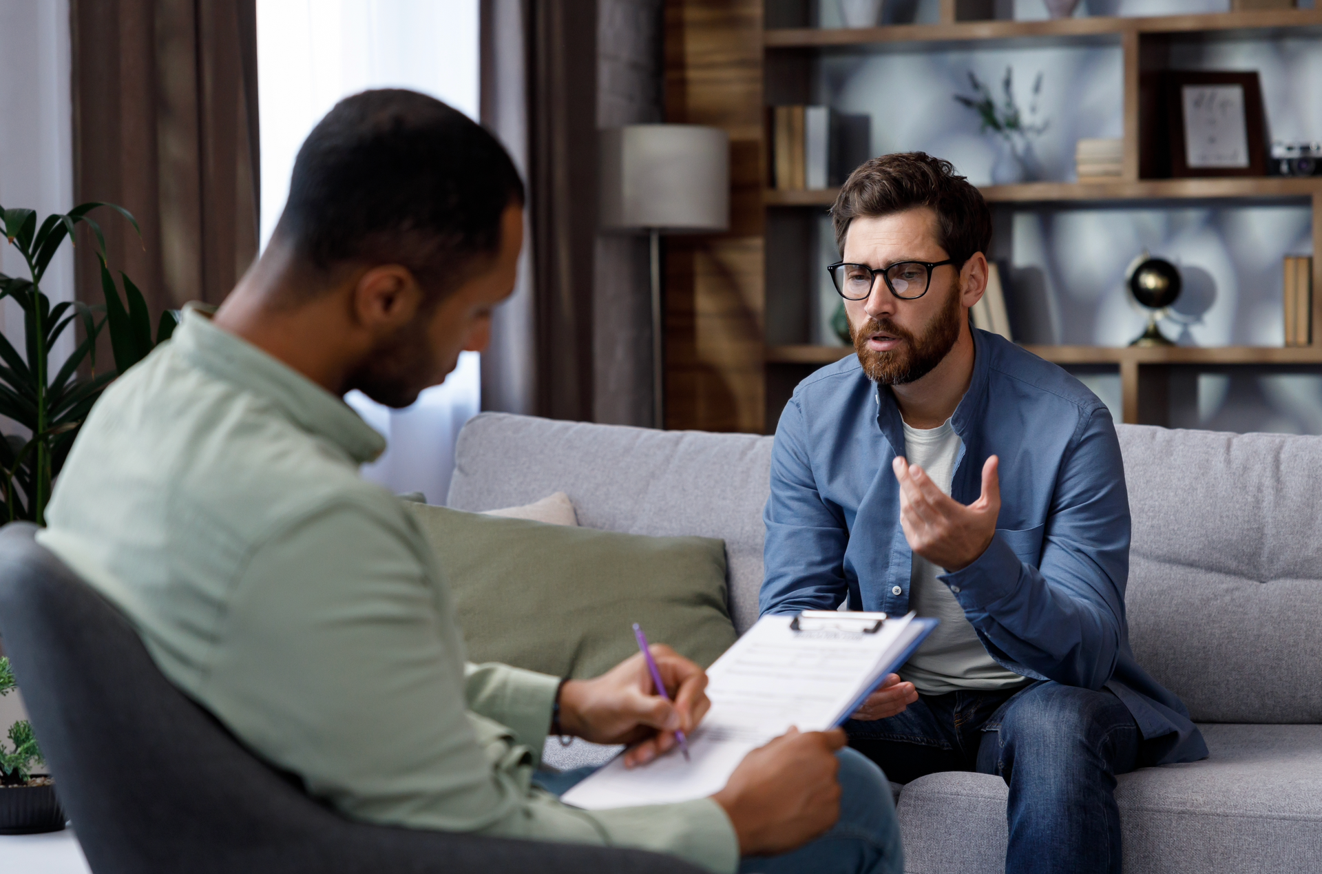 A man is sitting on a couch talking to another man while holding a clipboard.