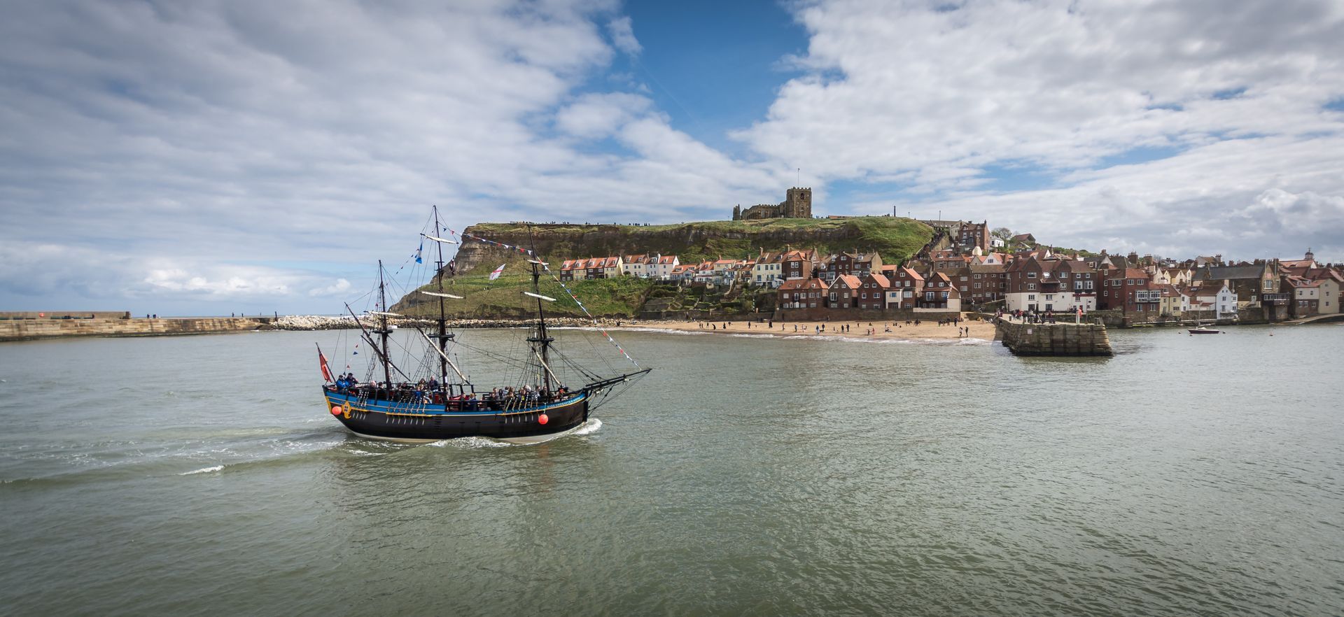 A black ship sails toward a coastal town with a castle on a hill, under a cloudy sky.
