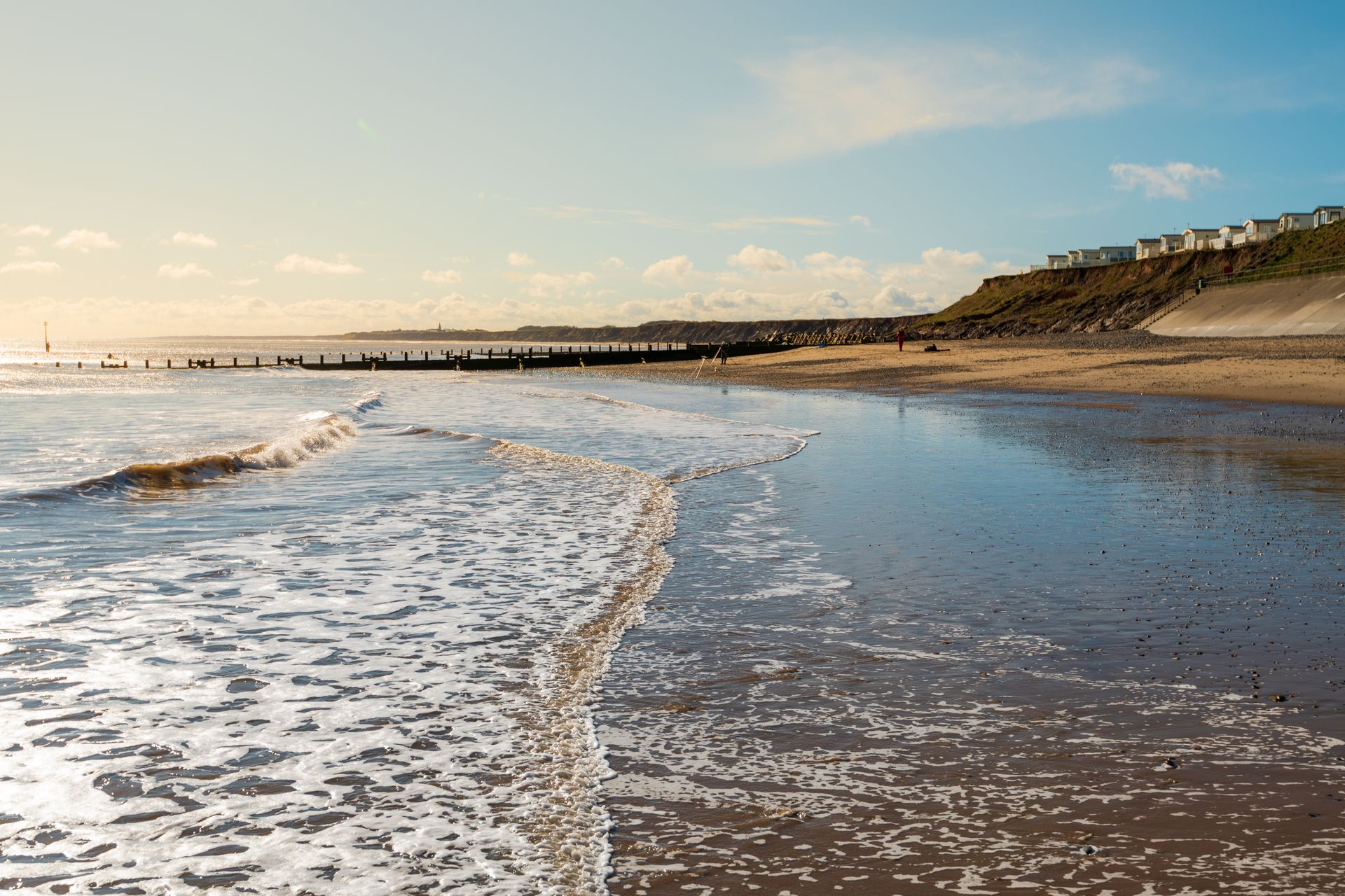 Waves wash over a sandy beach under a blue sky. Buildings sit atop a distant cliff.