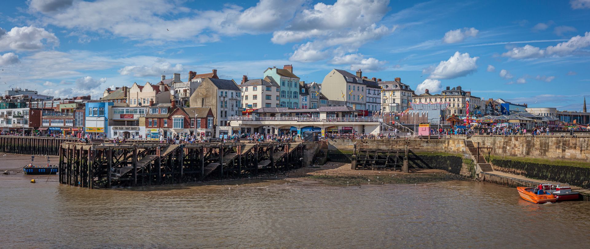 A coastal town with colorful buildings, people, and a water body with an orange boat under a cloudy blue sky.