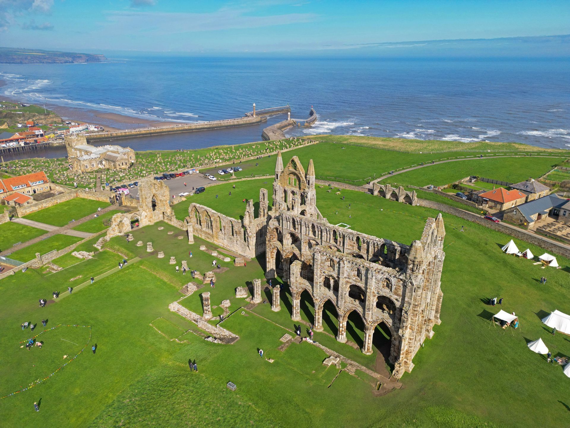 Whitby Abbey ruins on a green hill overlooking the ocean and a harbor on a sunny day.