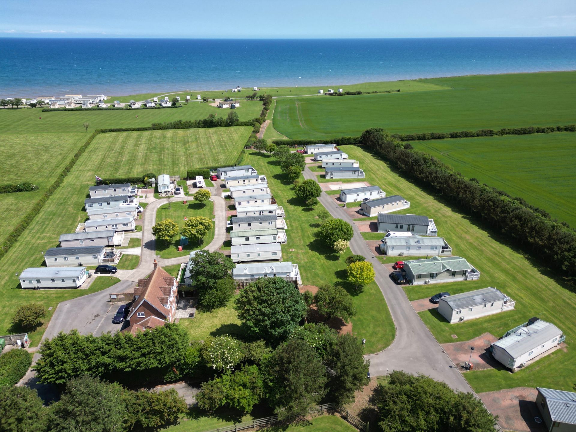 Aerial view of a caravan park near the sea. White caravans line grassy paths, green fields and blue ocean in background.