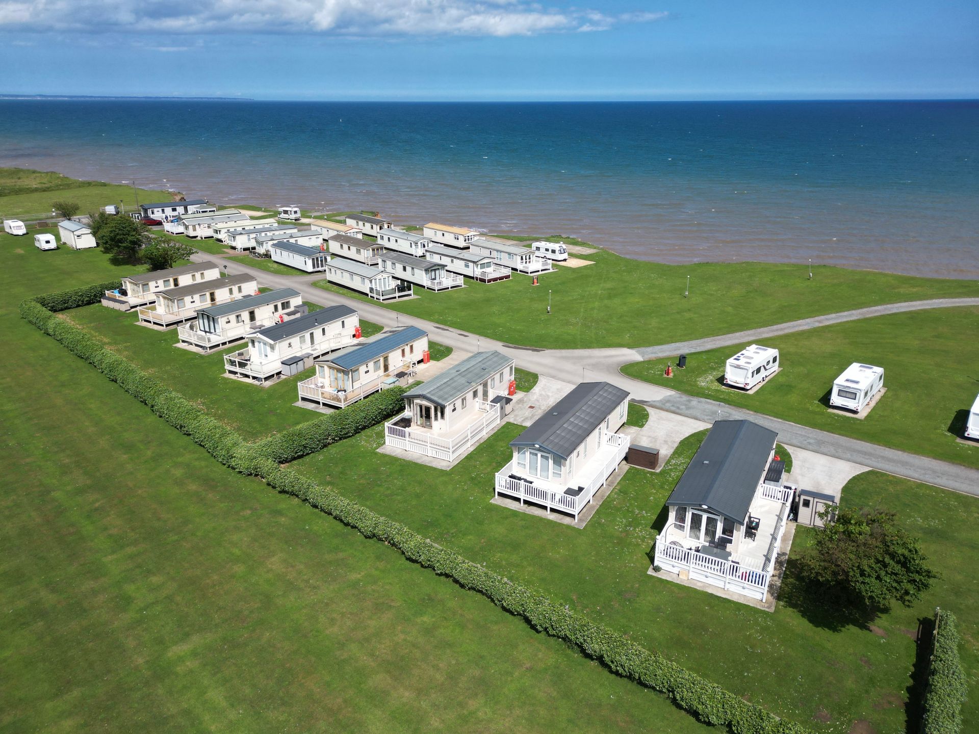 Aerial view: caravan park on green grass beside a blue ocean under a sunny sky.