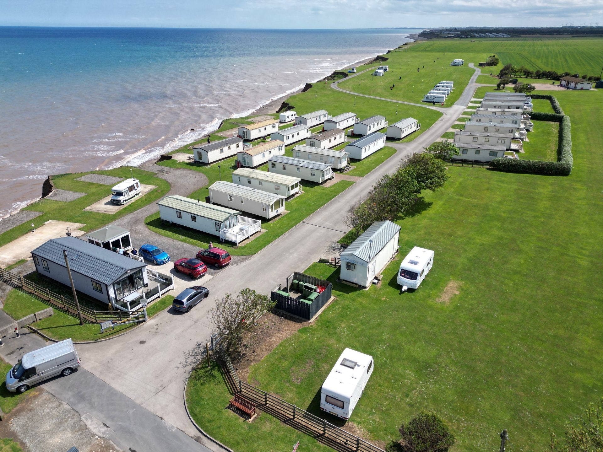 Aerial view of a seaside caravan park with white caravans, green grass, and the blue ocean.