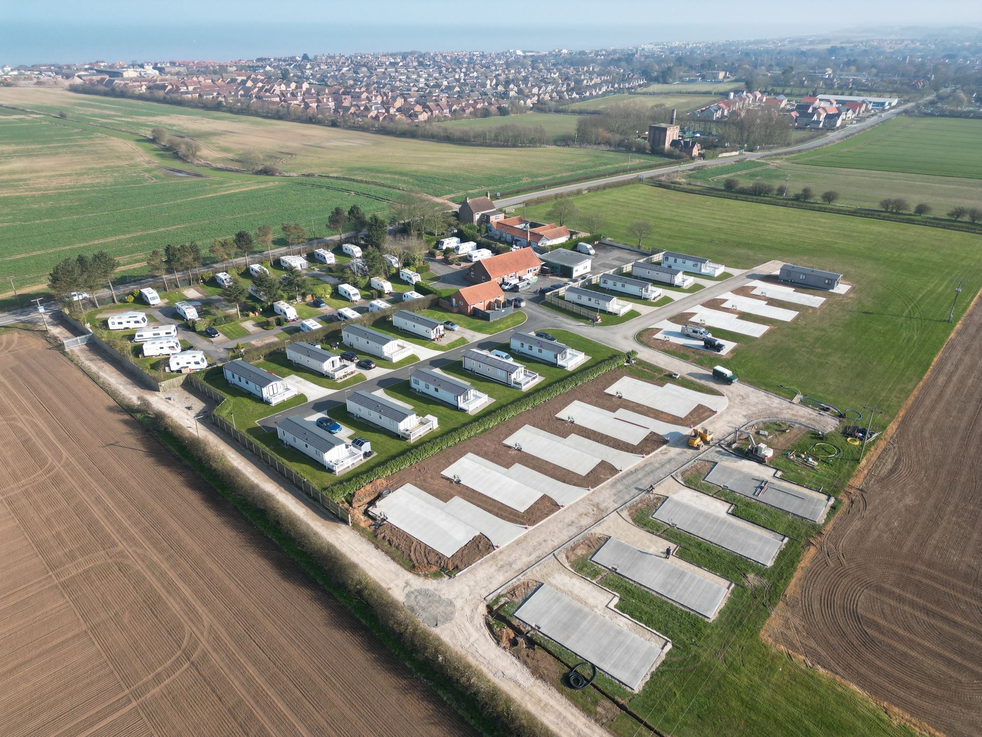 Aerial view of a caravan park with rows of white caravans, green fields, and a town in the distance.