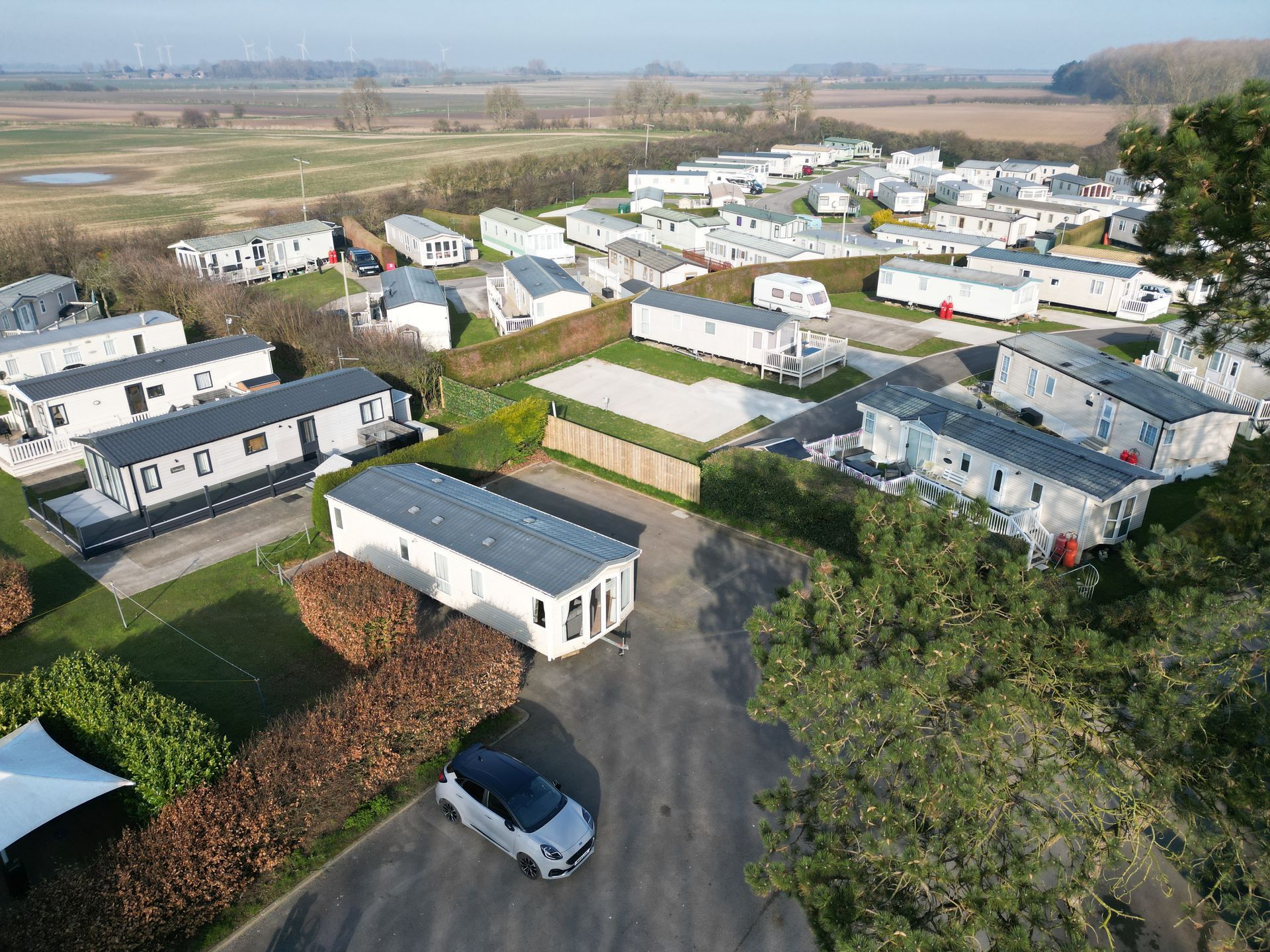 Aerial view of a trailer park with many white and gray mobile homes; sunny day with greenery and a car.