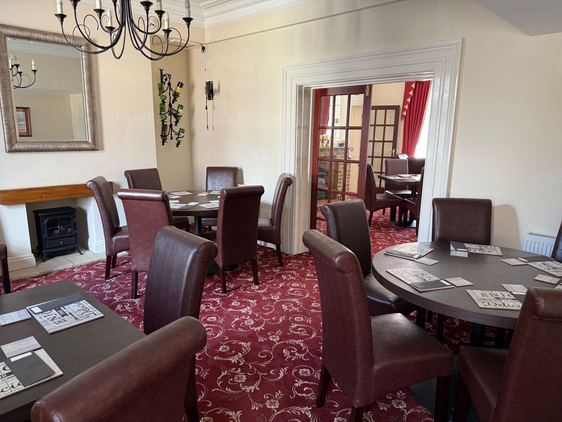 A room with round tables, burgundy chairs, and a red patterned carpet. A doorway leads to another room.