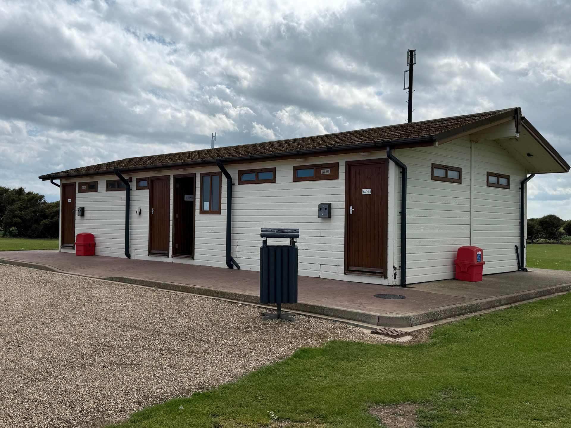 Public restroom building with white siding, brown doors, red bins, and a dark gray trash can on gravel.