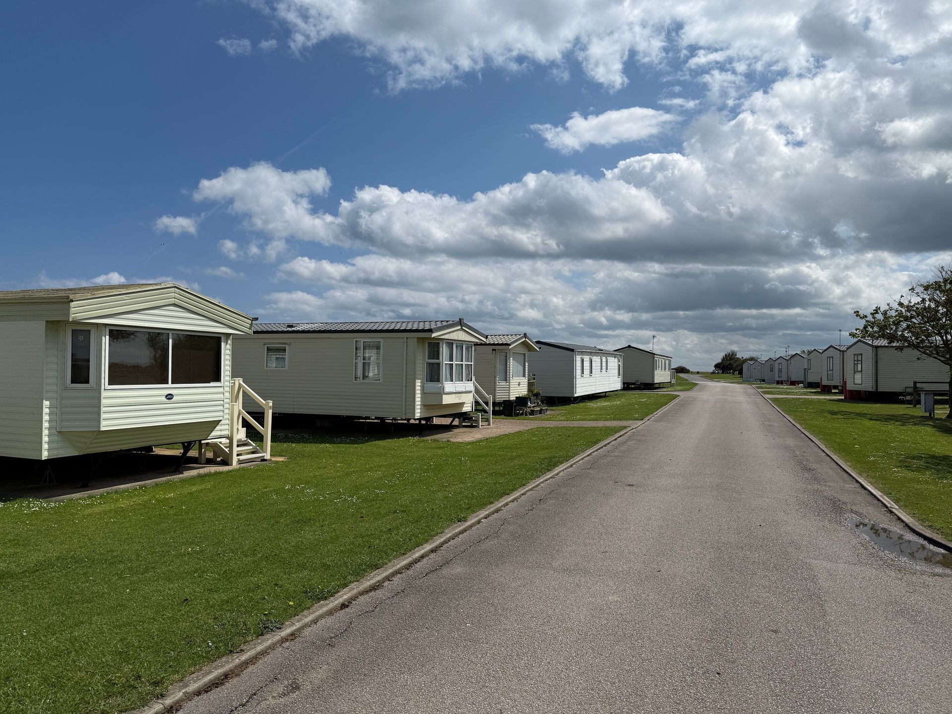 Row of white mobile homes on a green lawn, with a paved road under a partly cloudy blue sky.