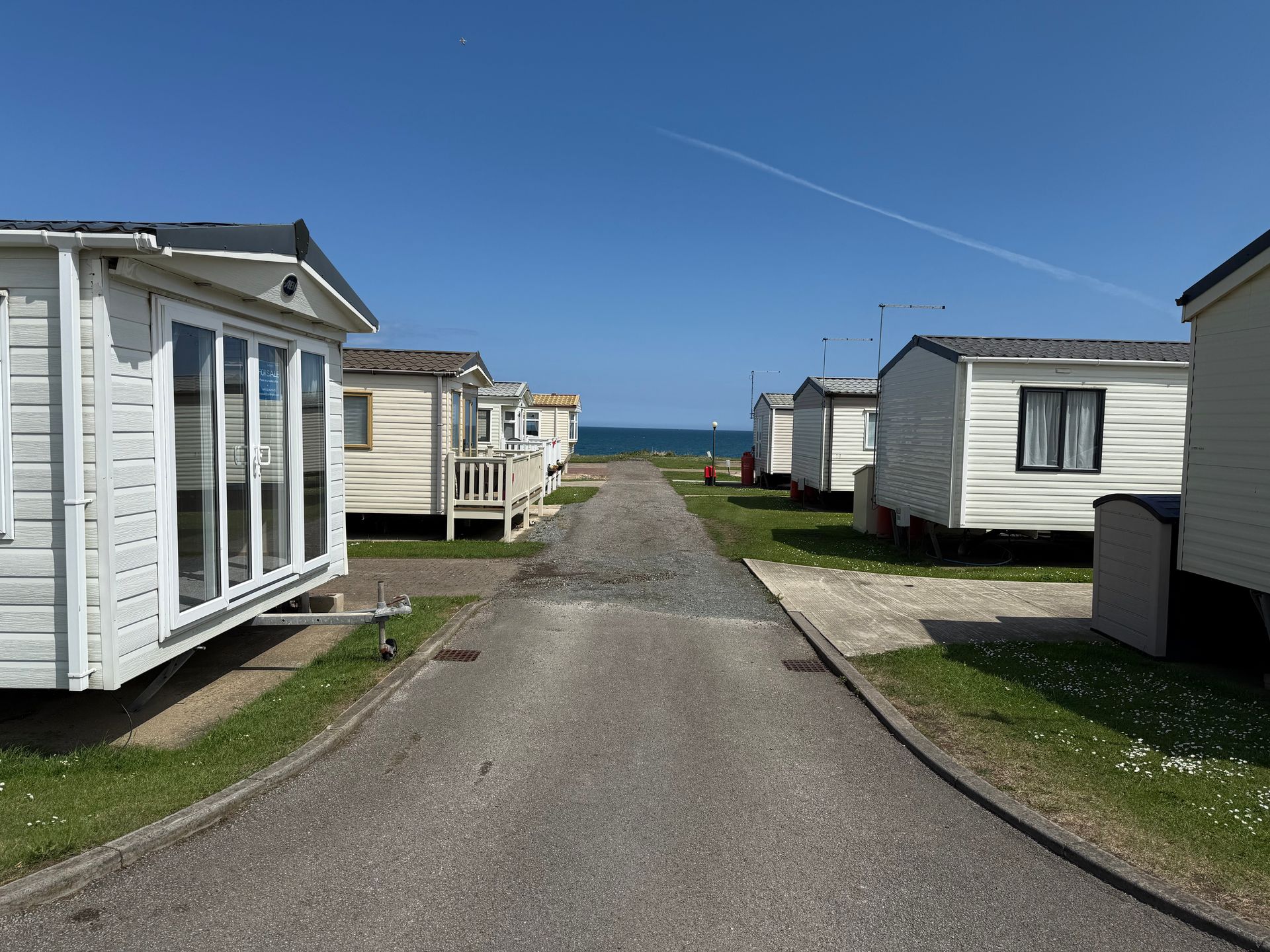 A gravel road between white mobile homes leads to a blue sea under a cloudless sky.