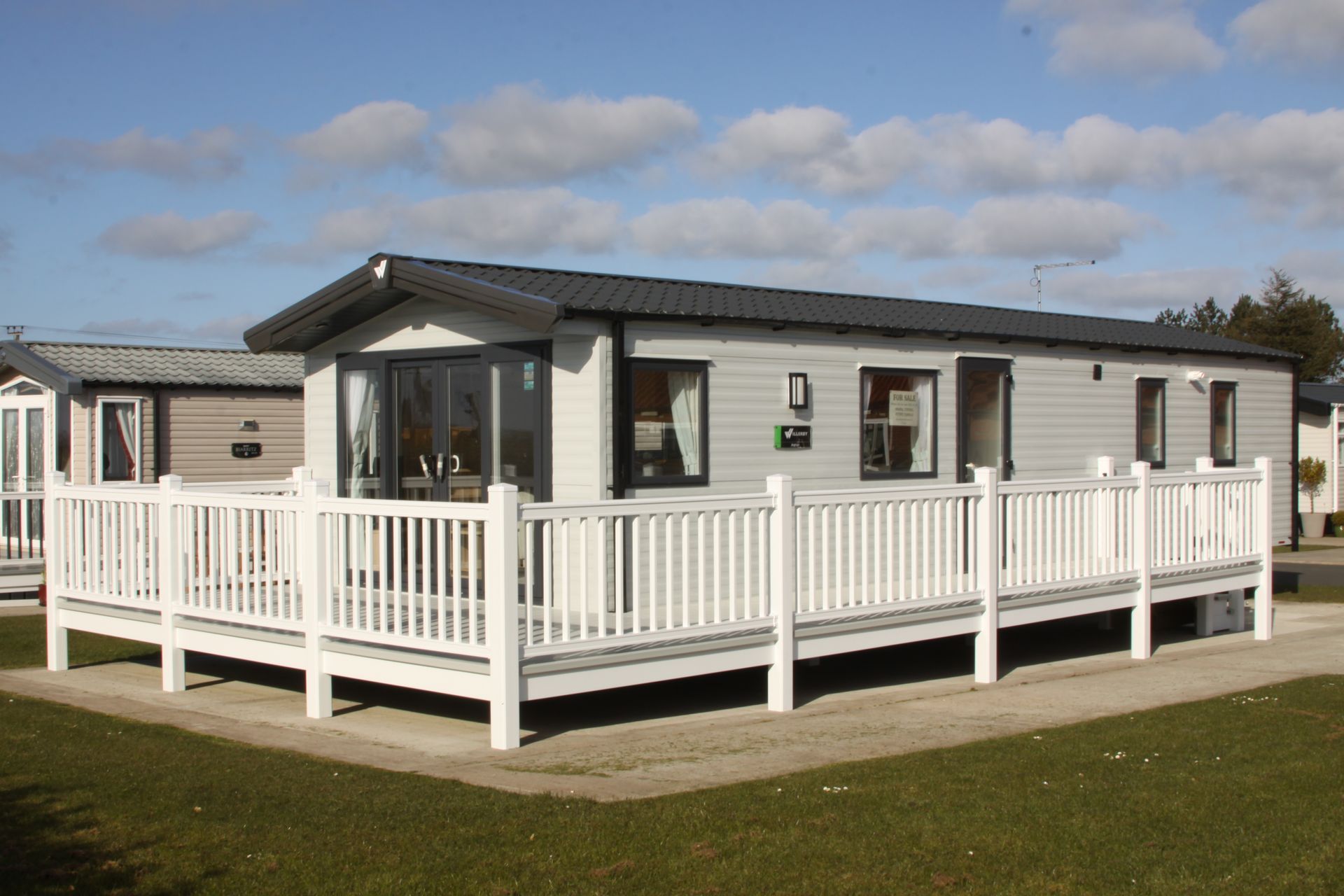 Gray static caravan with a white deck on a grassy lot, under a partly cloudy sky.