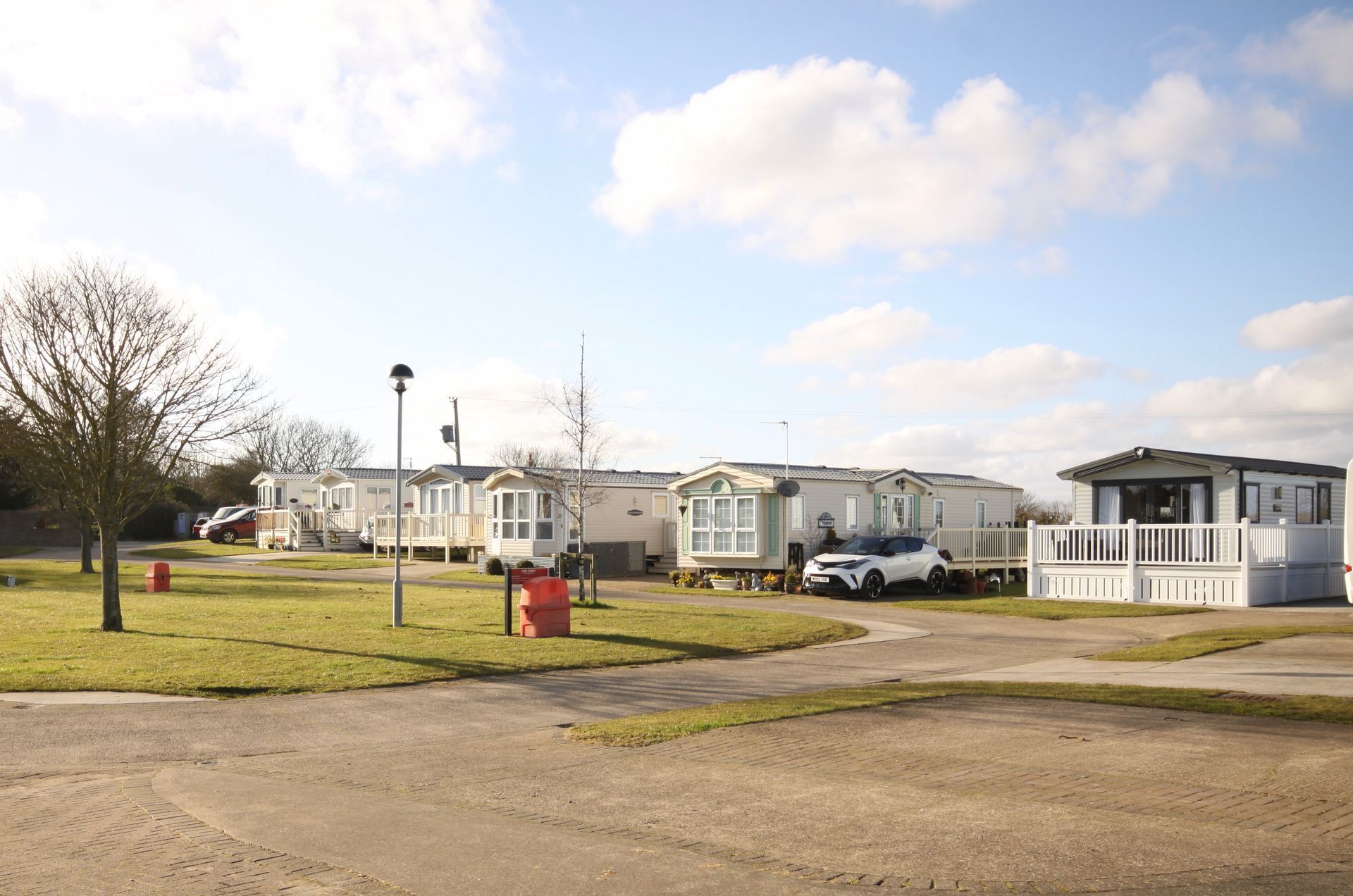 Mobile homes in a grassy area with a cloudy blue sky. A car is parked in front of one.