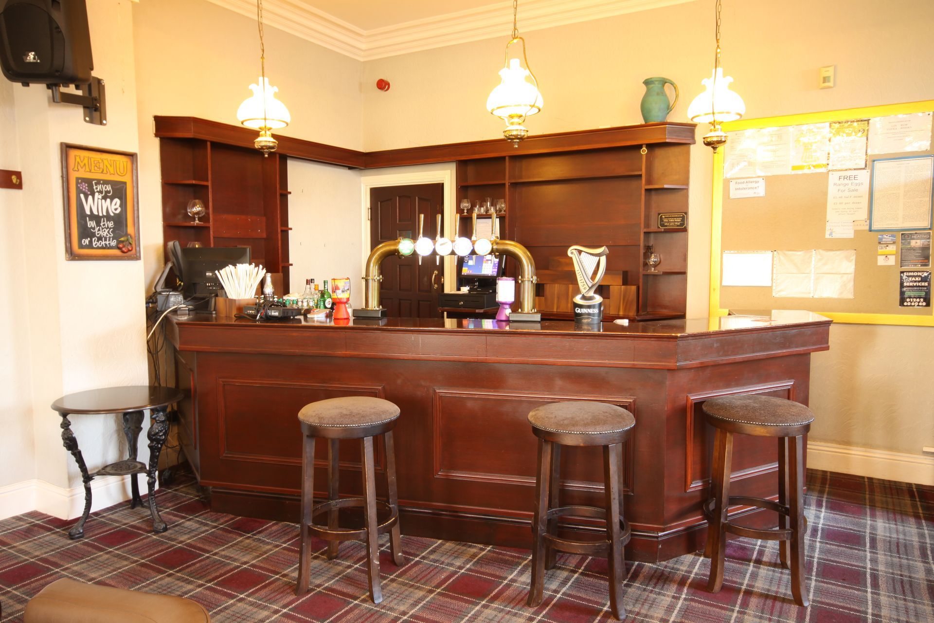 A wooden bar with stools inside, with a tap and shelves. A corkboard is to the right.