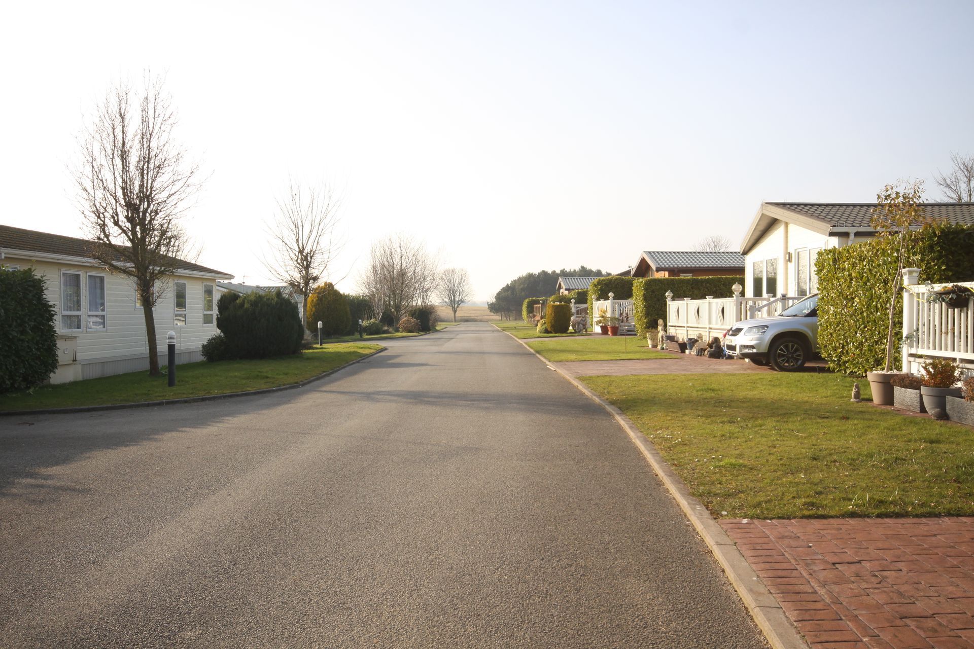 An asphalt road through a trailer park. White trailers with green lawns line the road. Bright sky.
