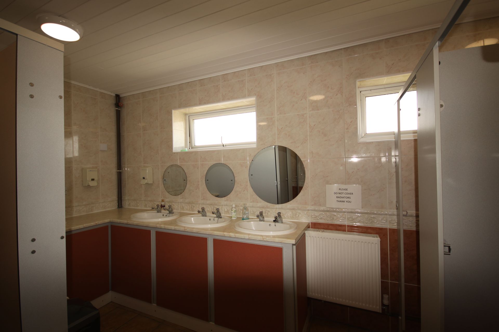 Public restroom with three sinks, three round mirrors, and two small windows. Red cabinets, white walls.