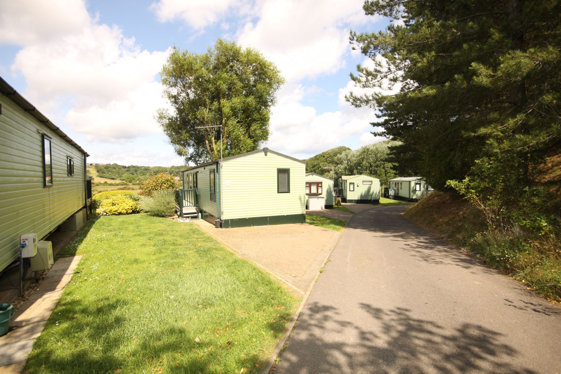 Mobile homes on a paved road, surrounded by grass, trees, and bright sunny sky.