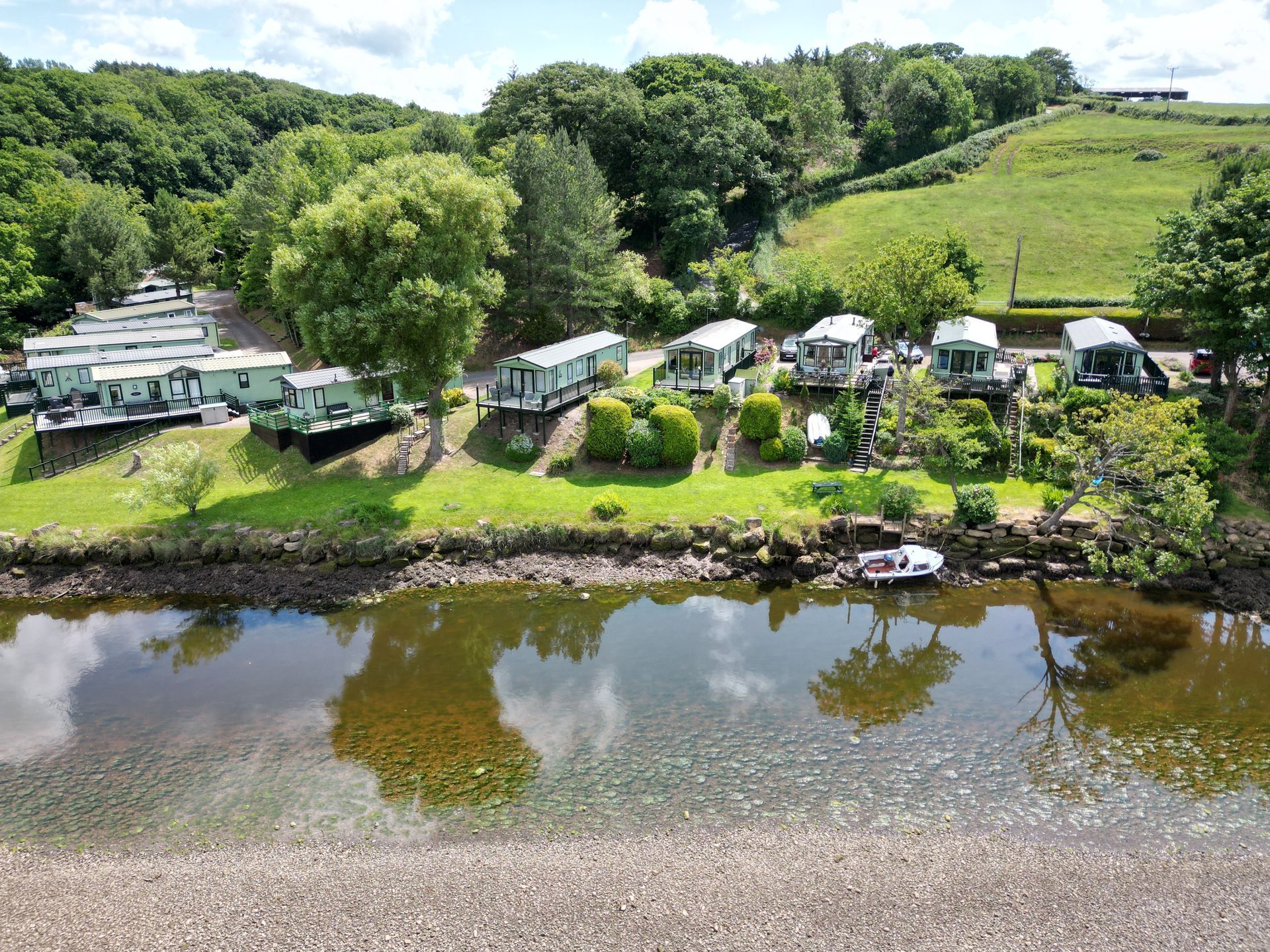 Campground along river with trailers, green grass, trees, and a hillside.