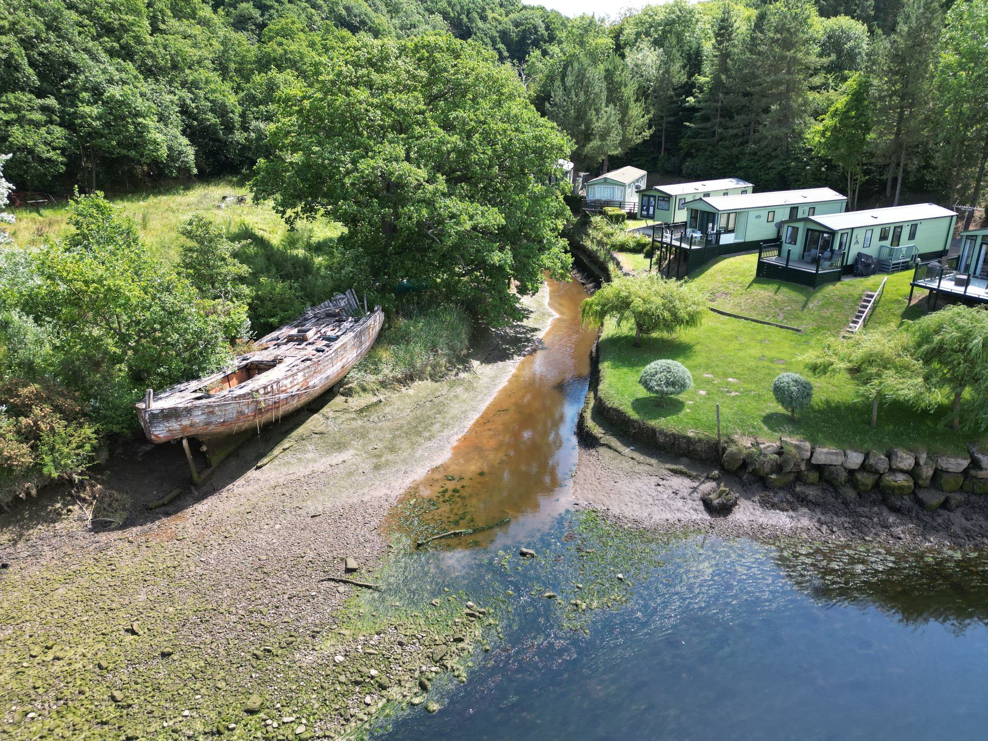 Derelict boat on muddy shore; mobile homes and trees in the background. Overcast day.