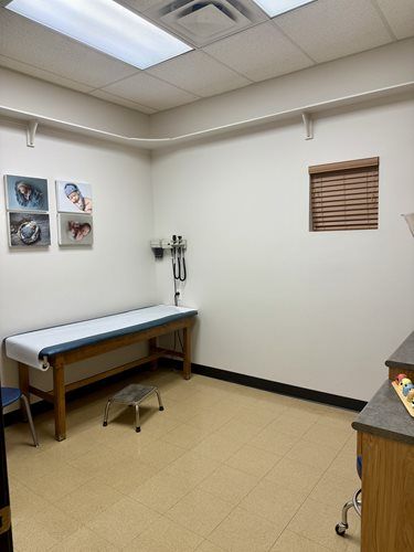 A clean doctor's examination room with a patient bed, a footstool, wall art, and medical equipment on the wall.