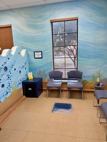 A waiting area with blue wave-themed wall murals, two grey chairs, a small dark table, and a window with blinds.