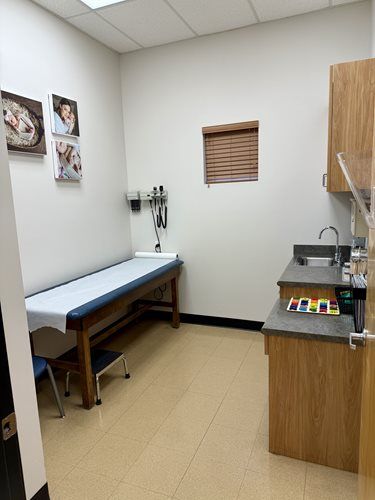 A clean medical examination room with an exam table, medical wall equipment, and a small sink area with light-tiled floors.