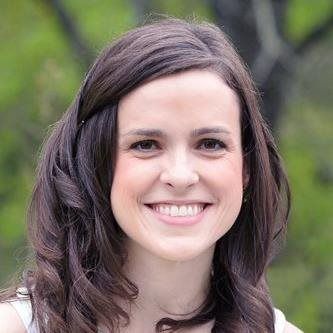 A smiling person with long, dark, wavy hair, shown from the chest up against a blurred green outdoor background.