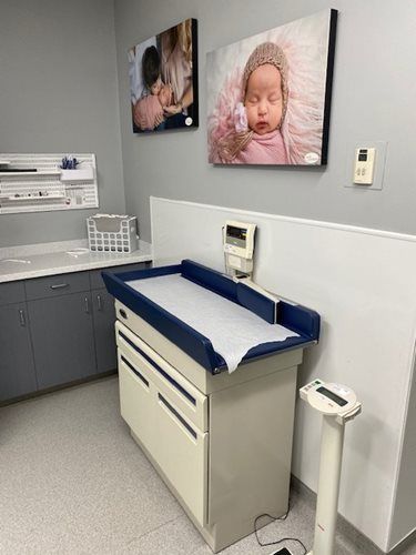 A pediatrician’s examination room with a white changing table, infant scale, and two framed photos of babies on the wall.