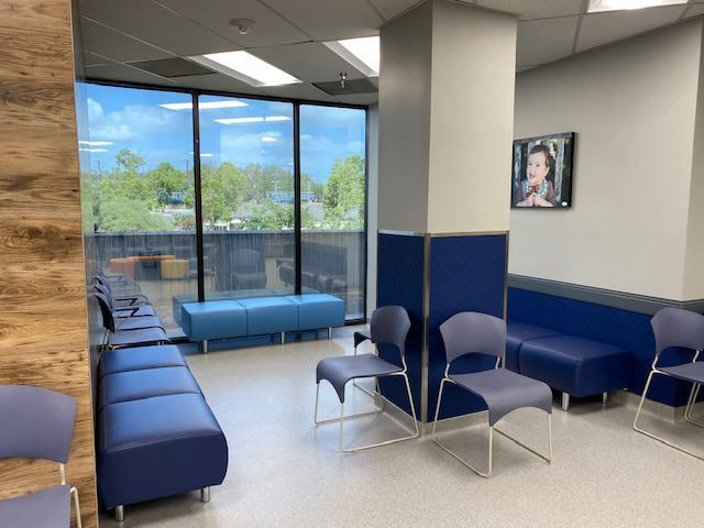 A modern medical waiting room with blue seating, chairs, a structural column, and a large window looking onto trees.