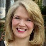 A headshot of a smiling person with shoulder-length blonde hair, wearing a dark top, set against a blurred background.