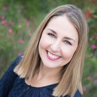 A woman smiling at the camera, wearing a dark blue top against a soft-focus, natural background of greenery and flowers.