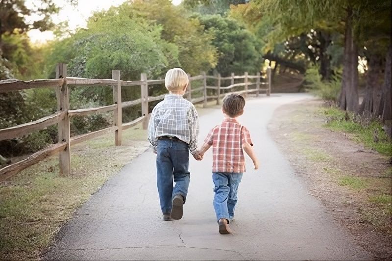 Two children holding hands while walking down a tree-lined path beside a wooden fence.