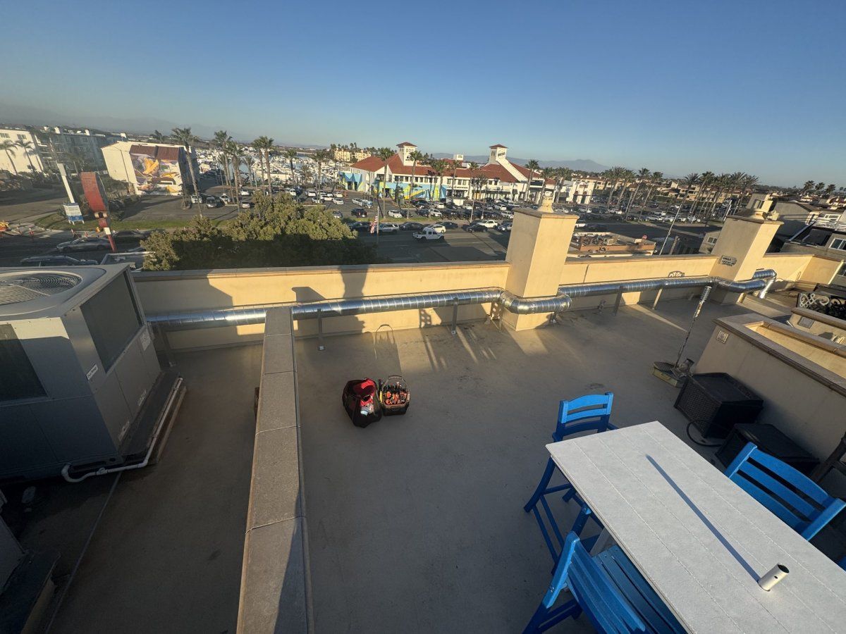 Rooftop view of a small robot vacuuming. Blue table and chairs on the right. City in background under blue sky.